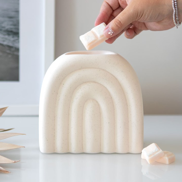 Hand dipping a small rectangular bar of soap into a beige, rainbow-shaped container on a white surface.