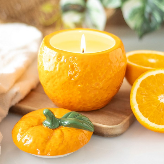 An orange-shaped ceramic candle holder with a lit candle, shown alongside a matching orange-shaped dish and sliced orange props on a wooden board.