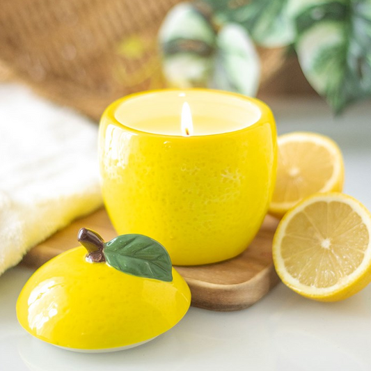 Yellow lemon-shaped candle with a lit wick on a white surface with lemons and leaves.