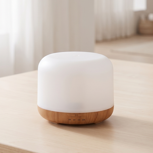 A small cylindrical essential oil diffuser with a white frosted top and a light wood base, sitting on a light wooden table in a softly lit room.