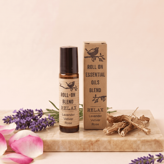 A roll-on essential oil bottle labeled 'RELAX Lavender Vetiver Rose' stands next to its kraft paper box on a marble slab, styled with lavender sprigs, pink petals, and dried roots against a soft beige background.