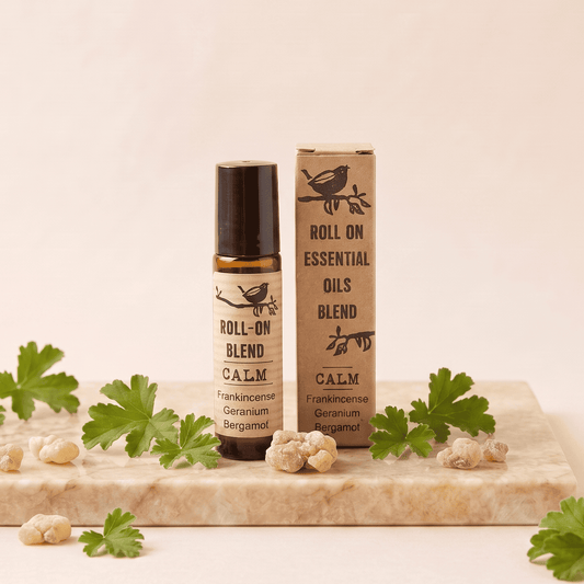 A roll-on essential oil blend bottle and its matching cardboard box, both labeled 'ROLL ON ESSENTIAL OILS OILS BLEND CALM', displayed on a light stone slab with green herb leaves and small dried botanical pieces around them.