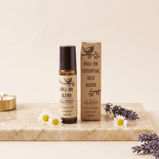 A roll-on essential oils blend bottle and its matching cardboard box, both labeled 'SLEEP Chamomile Lavender Herbal,' styled on a marble board with chamomile flowers, lavender sprigs, and a small candle against a warm cream background.