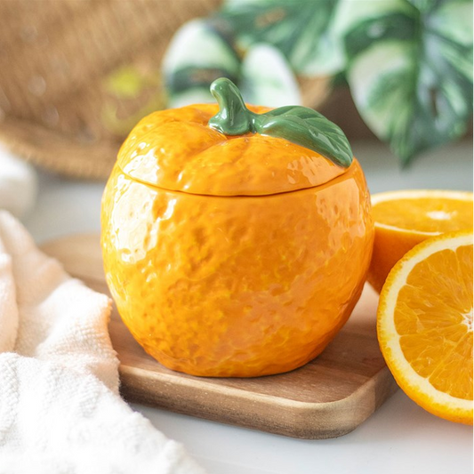 A ceramic orange-shaped container with a lid and a green stem, styled on a wooden board with sliced oranges and a white cloth in the background.