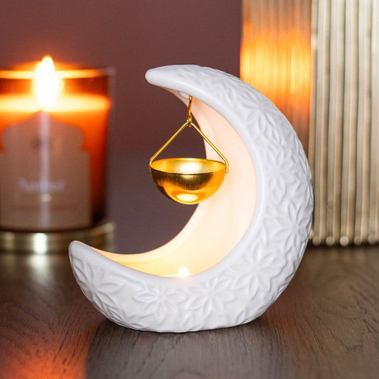 White ceramic moon-shaped lantern with a candle inside, placed on a wooden surface with a blurred candle and lamp in the background.