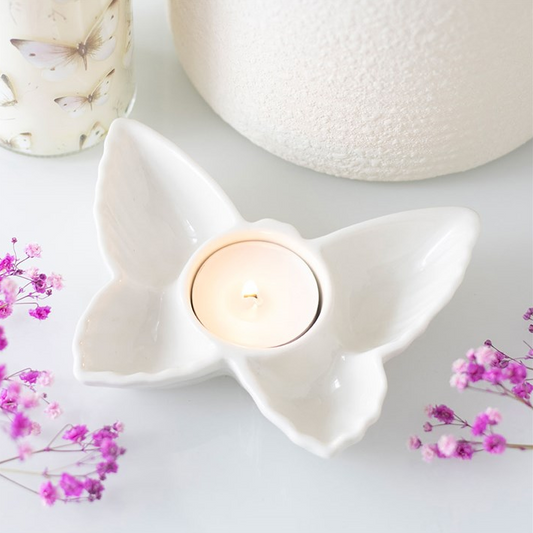 White ceramic butterfly-shaped candle holder with a lit candle on a light background with pink flowers.